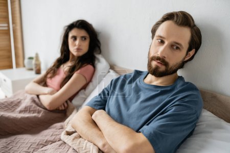 Nervous man crossing arms while sitting near blurred girlfriend on bed at home 