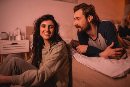 Bearded man talking to carefree girlfriend in bedroom in evening 