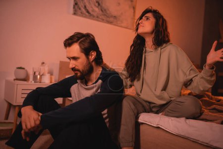Angry woman sitting near upset boyfriend in bedroom in evening 