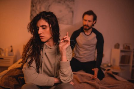 Sad brunette woman sitting near blurred boyfriend on bed at night 