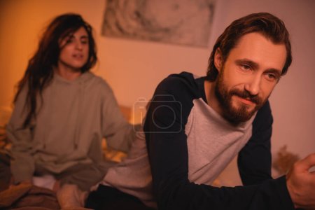 Indifferent man sitting near blurred girlfriend on bed in evening 