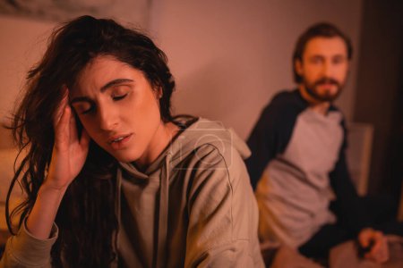 Dissatisfied woman touching head near blurred boyfriend at home 