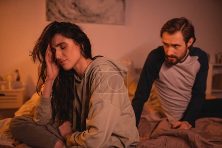 Upset woman sitting near blurred boyfriend talking on bed in evening 