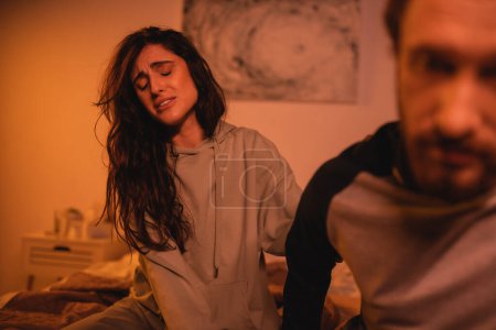 Dissatisfied woman sitting near blurred boyfriend in bedroom at night 