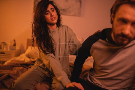 Upset brunette woman touching blurred boyfriend on bed at night 