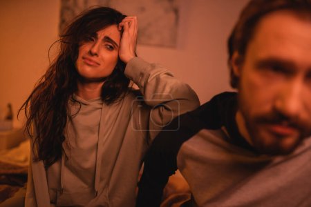 Displeased woman touching head and looking at blurred boyfriend in bedroom at night 