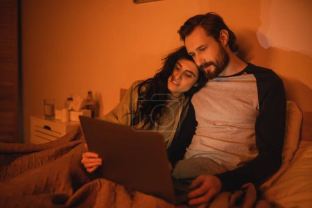 Smiling woman looking at laptop near bearded boyfriend on bed at night 