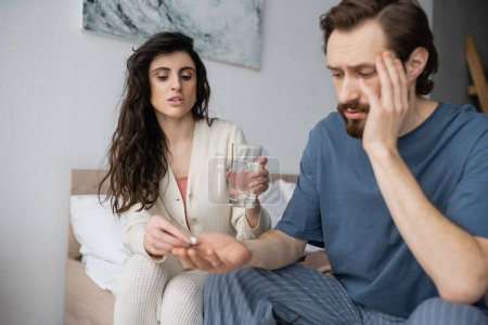 Caring woman giving pill and holding water near blurred boyfriend in bedroom at home