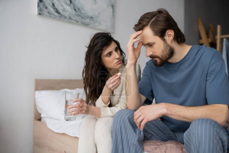 Caring woman holding pill and glass of water near boyfriend suffering from migraine in bedroom  