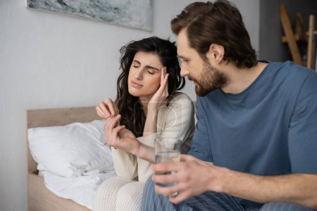 Caring man giving pills and water to girlfriend with migraine in bedroom 