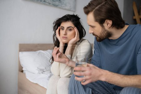 Caring man holding pills and water near frustrated girlfriend on bed at home 