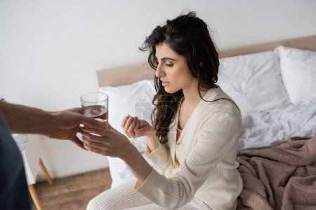 Caring man giving water to girlfriend with pill sitting on bed in morning 