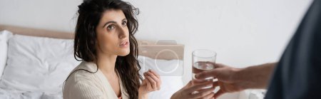 Foto de Mujer morena sosteniendo pastillas y tomando un vaso de agua de novio borroso en el dormitorio, pancarta - Imagen libre de derechos