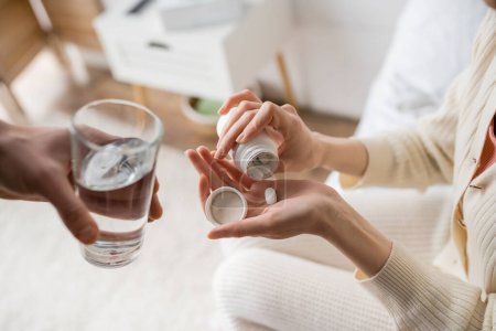 Vista recortada de la mujer vertiendo píldora en la mano cerca de novio con vaso de agua en casa 