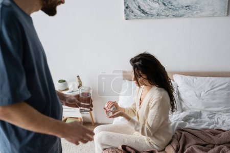 Woman holding pills near blurred boyfriend with glass of water in bedroom 