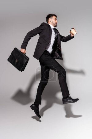 Foto de Vista lateral de hombre de negocios con estilo en traje negro levitando con café para llevar y maletín de cuero sobre fondo gris - Imagen libre de derechos