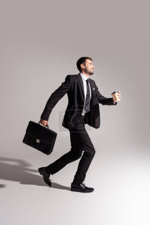 Foto de Vista lateral del hombre de negocios sonriente con bebida para llevar y maletín corriendo sobre fondo gris - Imagen libre de derechos