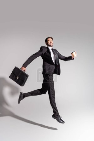Foto de Vista lateral del hombre de negocios encantado con taza desechable y maletín levitando sobre fondo gris - Imagen libre de derechos