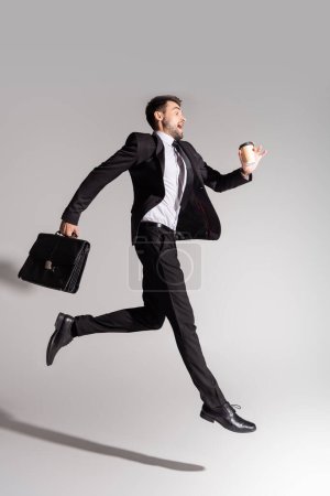Foto de Vista lateral del hombre de negocios emocionado en traje negro levitando con maletín de cuero y taza de papel sobre fondo gris - Imagen libre de derechos