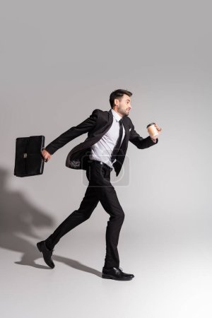 Photo for Side view of positive businessman in black suit running with briefcase and paper cup on grey background - Royalty Free Image