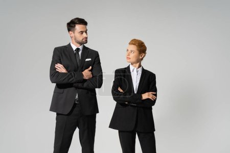 skeptical business people in black pantsuits standing with folded arms and looking at each other isolated on grey