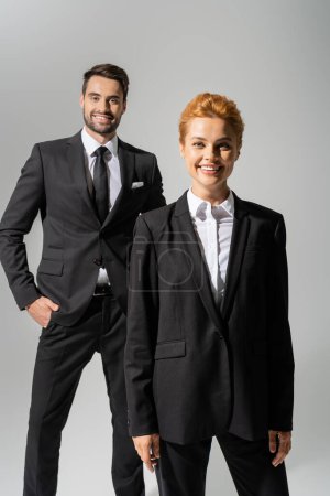 Photo for Cheerful redhead businesswoman looking at camera near smiling colleague standing with hand in pocket isolated on grey - Royalty Free Image