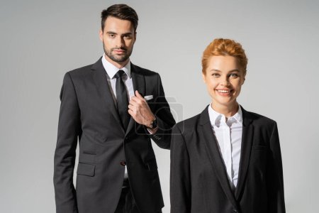 Photo for Cheerful redhead businesswoman smiling at camera near confident businessman in stylish suit isolated on grey - Royalty Free Image