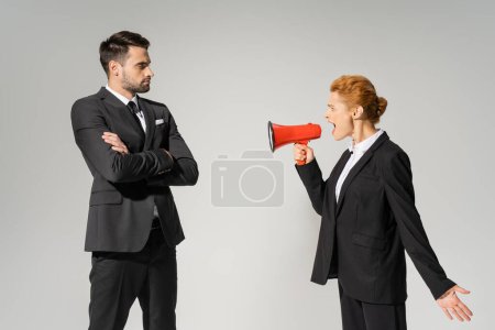 angry businesswoman screaming in megaphone at confident man in suit standing with crossed arms isolated on grey