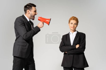 hombre de negocios enojado gritando en altavoz cerca de mujer escéptica de pie con los brazos cruzados aislados en gris