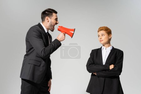 angry boss screaming in megaphone near indifferent redhead employee standing with folded arms isolated on grey