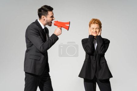 Téléchargez les photos : Gérant rousse avec les yeux fermés couvrant les oreilles avec les mains près du patron criant dans le mégaphone isolé sur gris - en image libre de droit