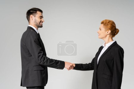 Photo for Side view of joyful business partners in black blazers shaking hands isolated on grey - Royalty Free Image
