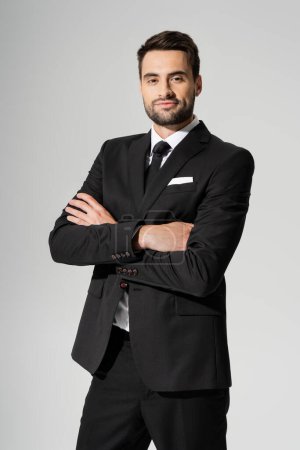 pleased bearded businessman in black stylish blazer crossing arms and looking at camera isolated on grey