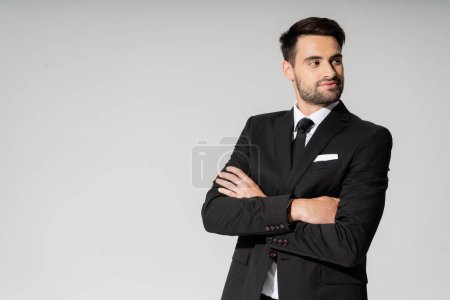 smiling businessman in black suit standing with folded arms and looking away isolated on grey