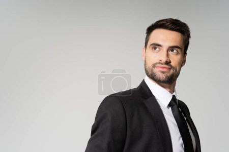 Photo for Smiling and dreamy businessman in black blazer looking away isolated on grey - Royalty Free Image