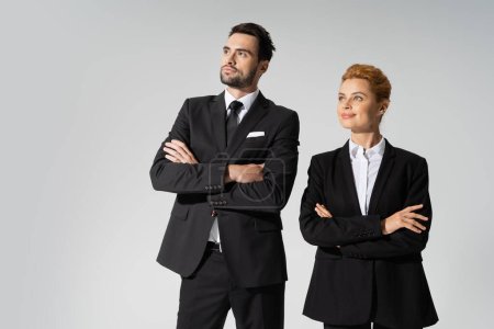 serious bearded businessman and cheerful redhead businesswoman crossing arms and looking away isolated on grey