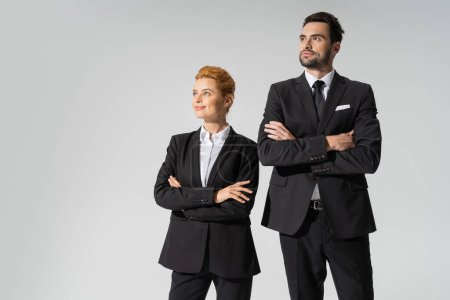 happy redhead businesswoman and confident businessman with crossed arms looking away isolated on grey