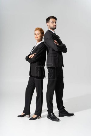 full length of confident redhead businesswoman standing back to back with business partner on grey background