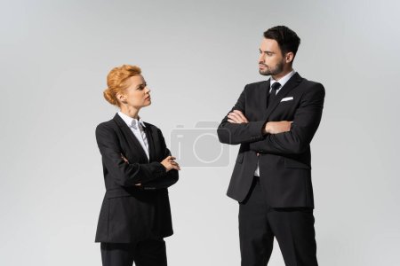 serious business people in black formal wear standing with crossed arms and looking at each other isolated on grey