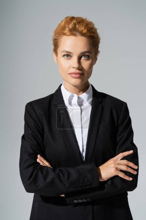 stylish redhead businesswoman in black blazer crossing arms and looking at camera isolated on grey