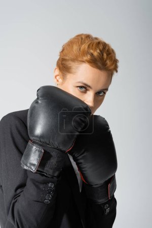 redhead businesswoman obscuring face with boxing gloves while looking at camera isolated on grey