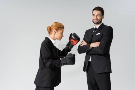 redhead woman in formal wear boxing near cheerful businessman standing with crossed arms isolated on grey