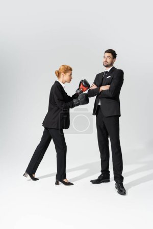 rothaarige Geschäftsfrau boxt neben lächelndem und skeptischem Geschäftsmann, der mit verschränkten Armen vor grauem Hintergrund steht