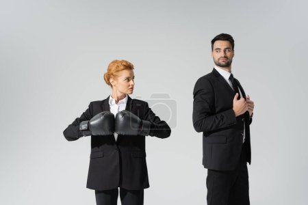 pelirroja mujer de negocios en guantes de boxeo mirando altivo hombre de negocios en chaqueta elegante aislado en gris