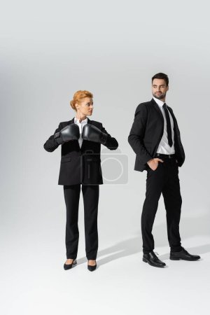 haughty businessman with hands in pockets looking at determined businesswoman in boxing gloves on grey background