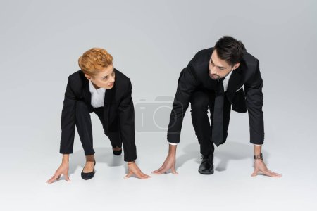 full length of business competitors in black formal wear standing in low start position and looking at each other on grey background