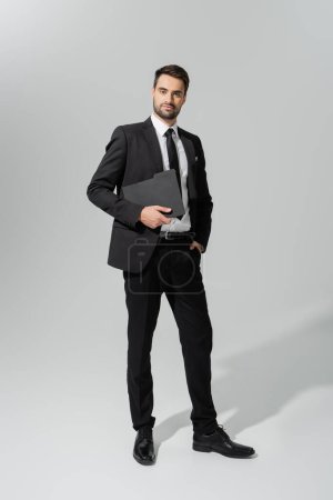 Téléchargez les photos : Pleine longueur de succès et confiant homme d'affaires en costume élégant noir debout avec dossier sur fond gris - en image libre de droit
