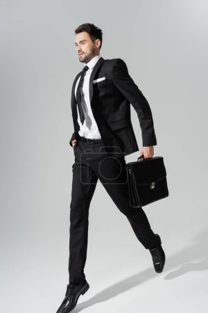 longitud completa de joven hombre de negocios en traje pantalón negro corriendo con maletín y mano en bolsillo sobre fondo gris