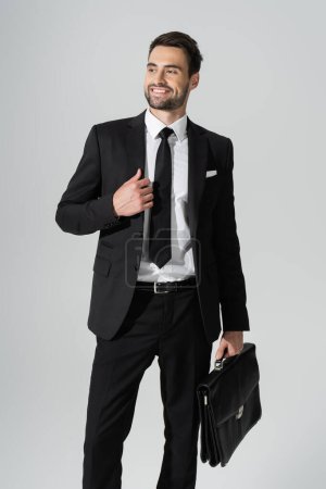 joyful businessman in black stylish pantsuit standing with briefcase and looking away isolated on grey