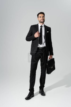 full length of happy successful businessman with leather briefcase looking away on grey background
